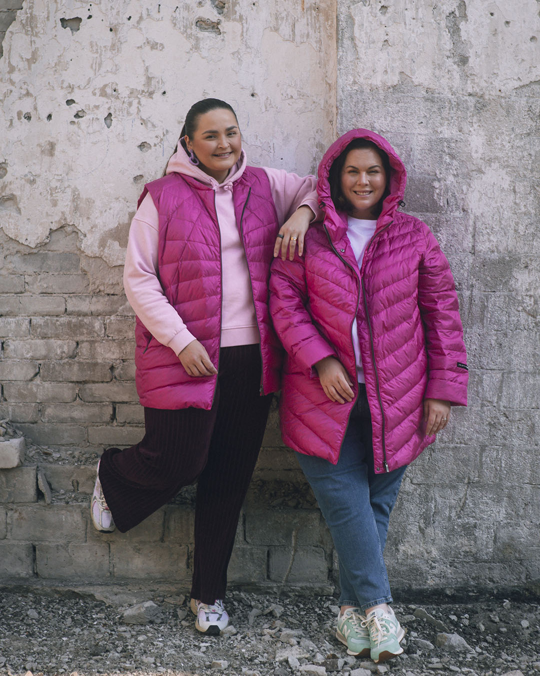 Two women are standing side by side against a rough stone wall. The person on the left is wearing a fuchsia Mesi down vest (color: Pitaya) and a light pink hoodie, while the one on the right is wearing a Jasmin light down coat (color: Pitaya) with a hood.