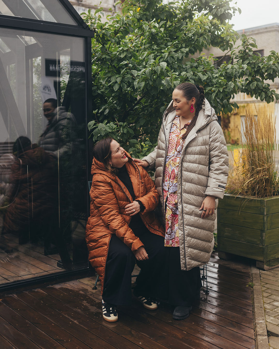 Two women are outside on a wooden terrace, one wearing a muted orange (color: rose gold) and the other a light (color: champagne) Säde light down coat. There is a colorful dress under the coat.