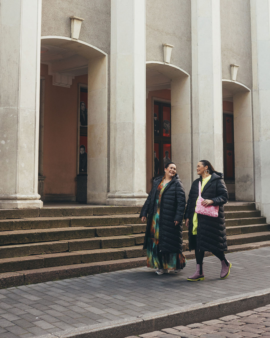 Two women are walking in the city wearing black Säde light down coats (color: charcoal black). Both coats are open, revealing colorful dresses and shoes underneath.