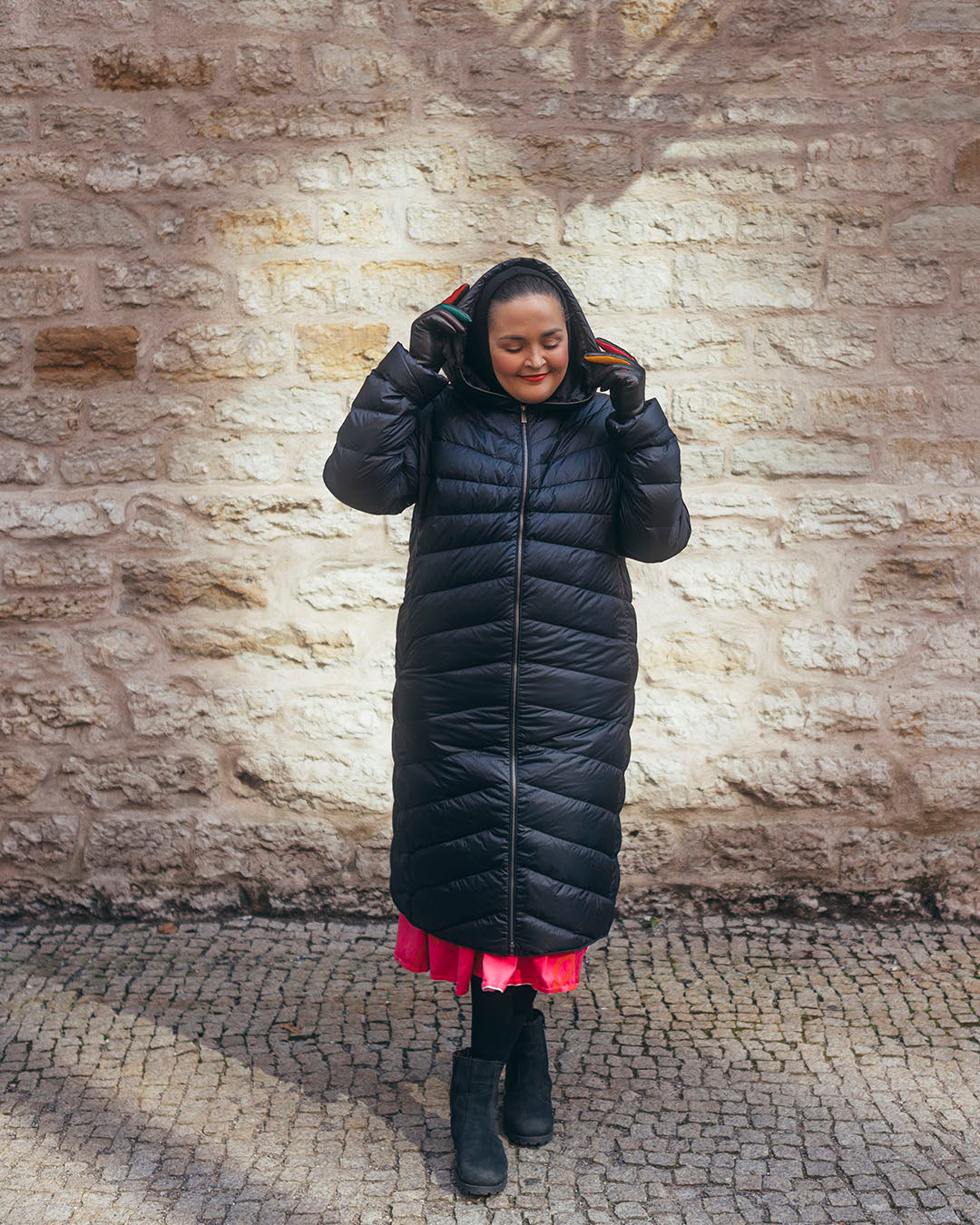 A woman is standing in front of a stone wall wearing a long black Säde light down coat (color: charcoal black) with the zipper closed and the hood up. A bright pink dress peeks out from underneath the coat.