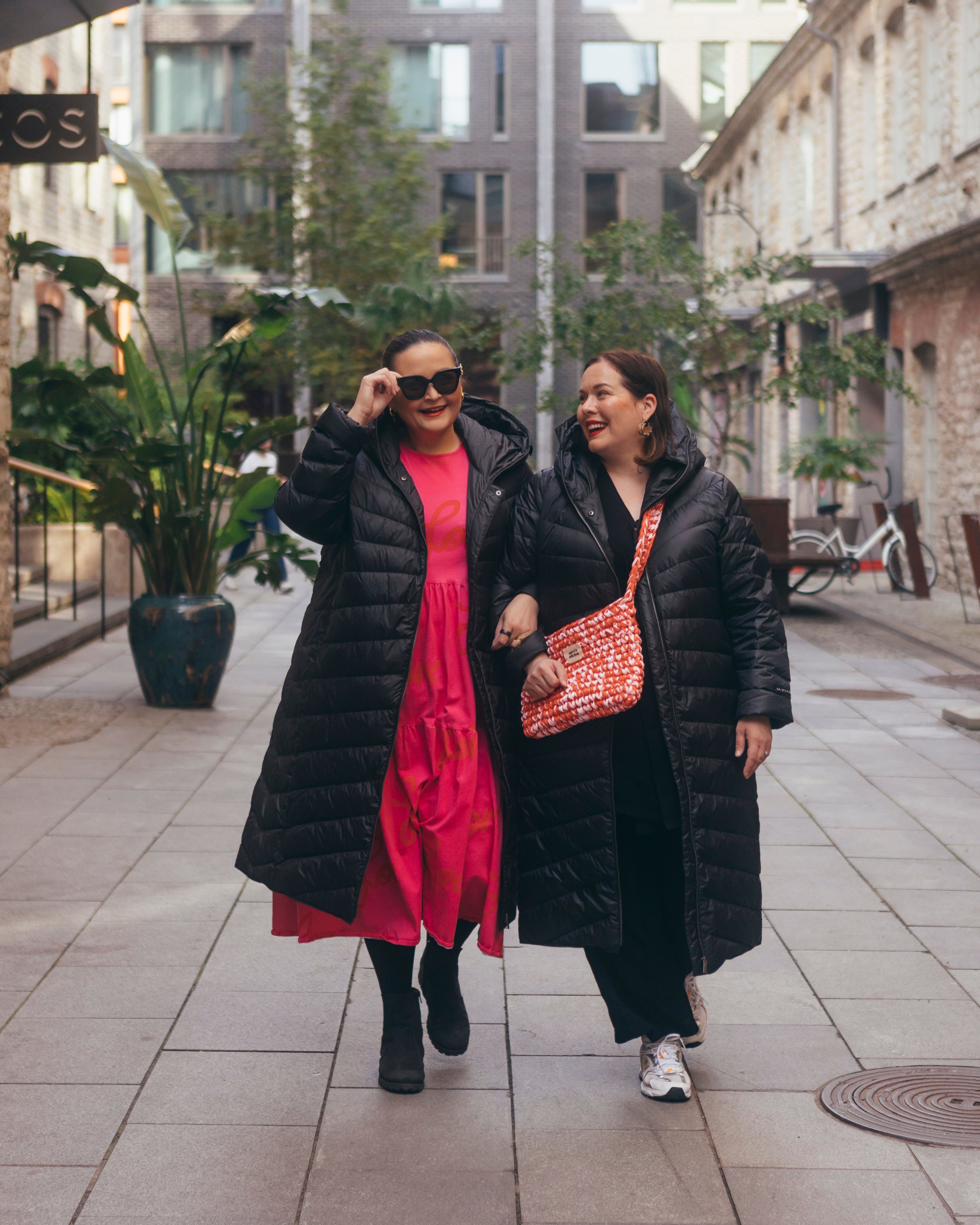 Two women are walking side by side in an urban street scene. Both are wearing long charcoal black Säde light down coats.