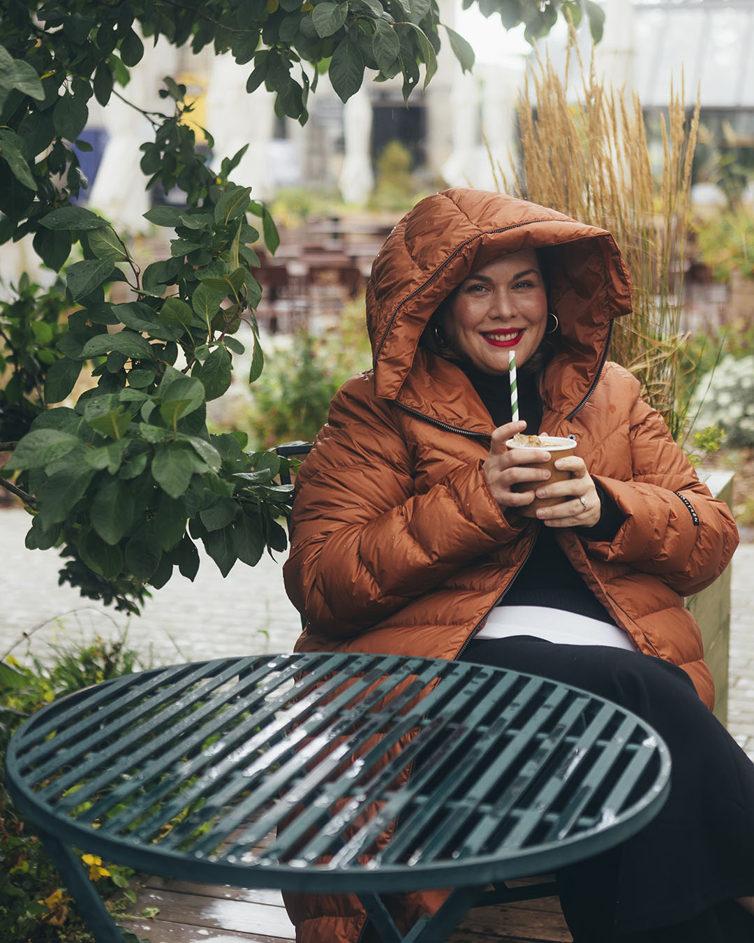 A woman is sitting outside smiling at a green table, drinking through a straw. She is wearing a muted orange Säde light down (color: rose gold) with the hood up.