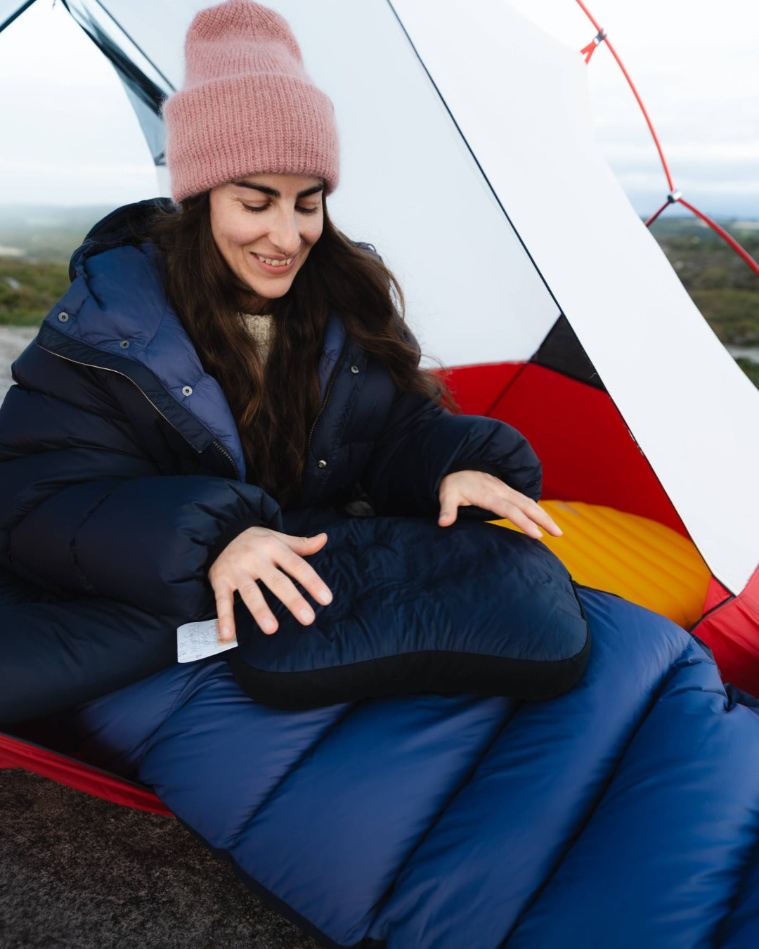 Woman sitting inside a sleeping bag in a tent, wearing a blue jacket and pink beanie, with a colorful interior.