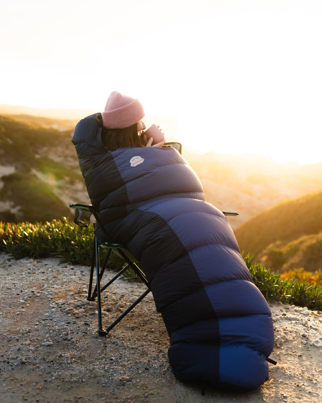 Person wrapped in a blue sleeping bag sitting on a chair with a scenic background