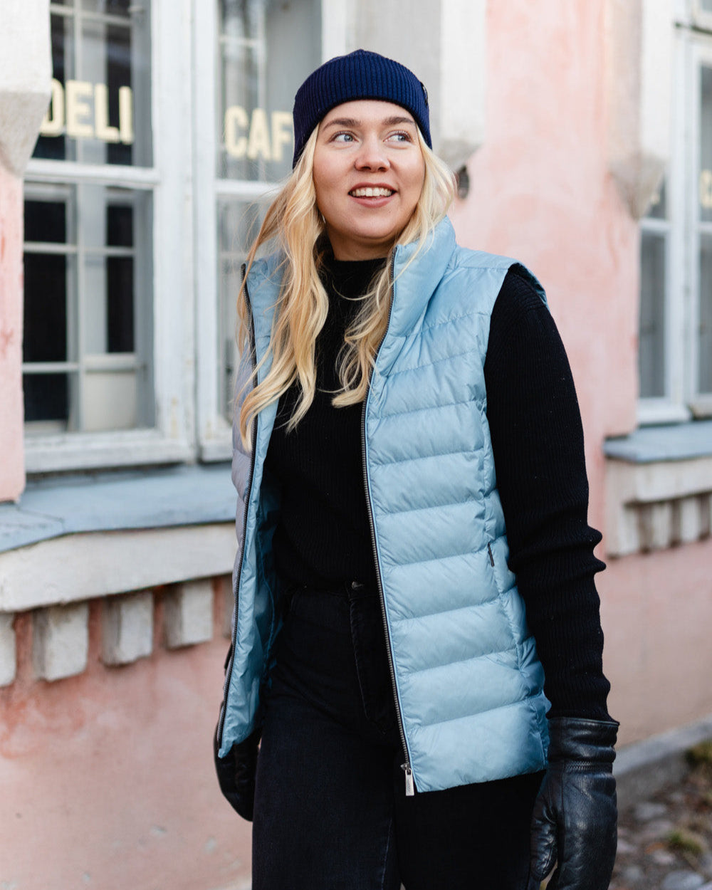 A woman stands on a street wearing a light blue Lady down vest (color: BlueBell). The vest has horizontal quilting and a slight sheen. She is also wearing a black beanie, a turtleneck sweater, trousers, and leather gloves.
