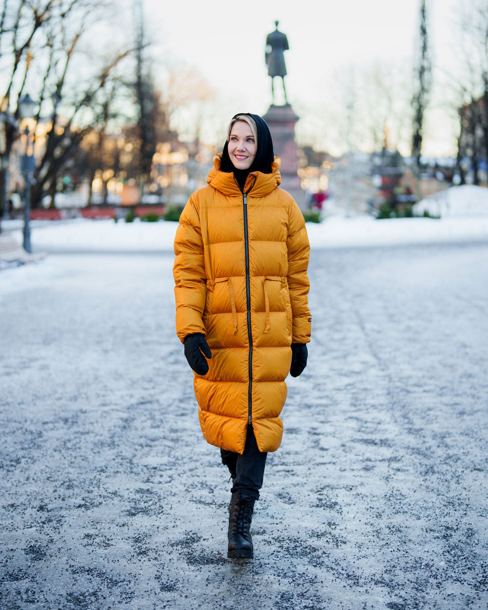 A smiling woman walks in a winter park wearing a yellow (color: Amber) Estrid down coat, black gloves, a black hood, and black boots. In the background, there is a snowy landscape and a statue