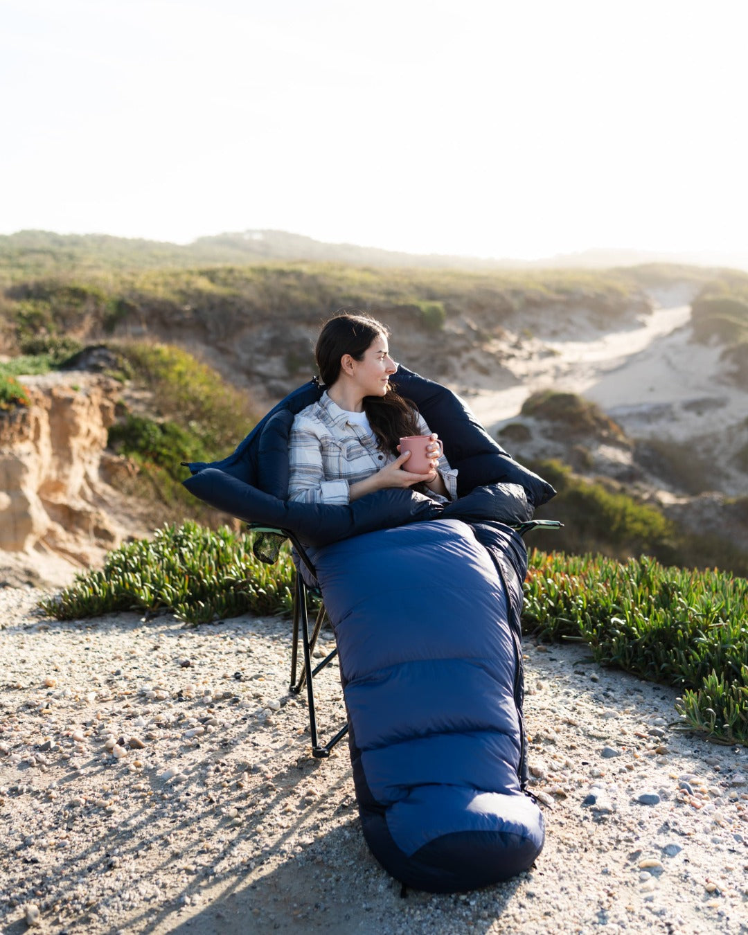 Person sitting on a beach with a sleeping bag, holding a cup