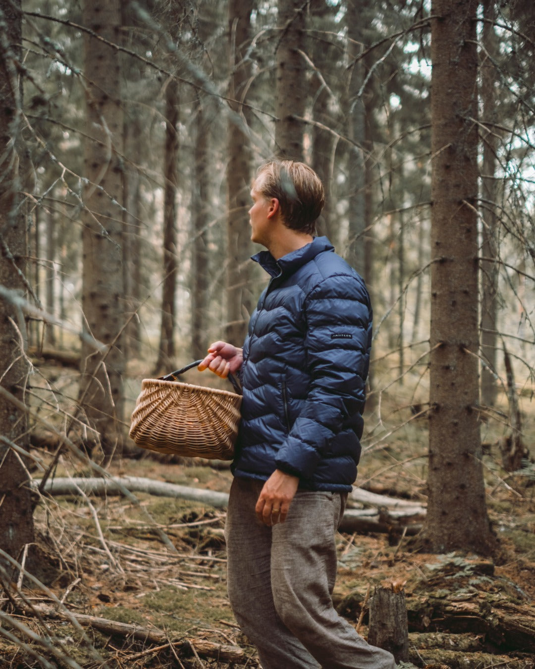 A man is standing in the forest holding a woven basket in his hand. He is wearing a blue Hile light down coat (color: Storm).