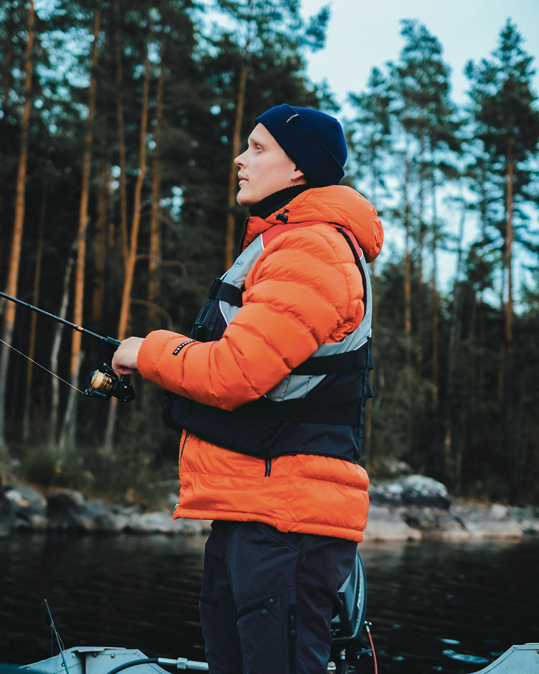 Man in an orange light down coat fishing by a forested lake