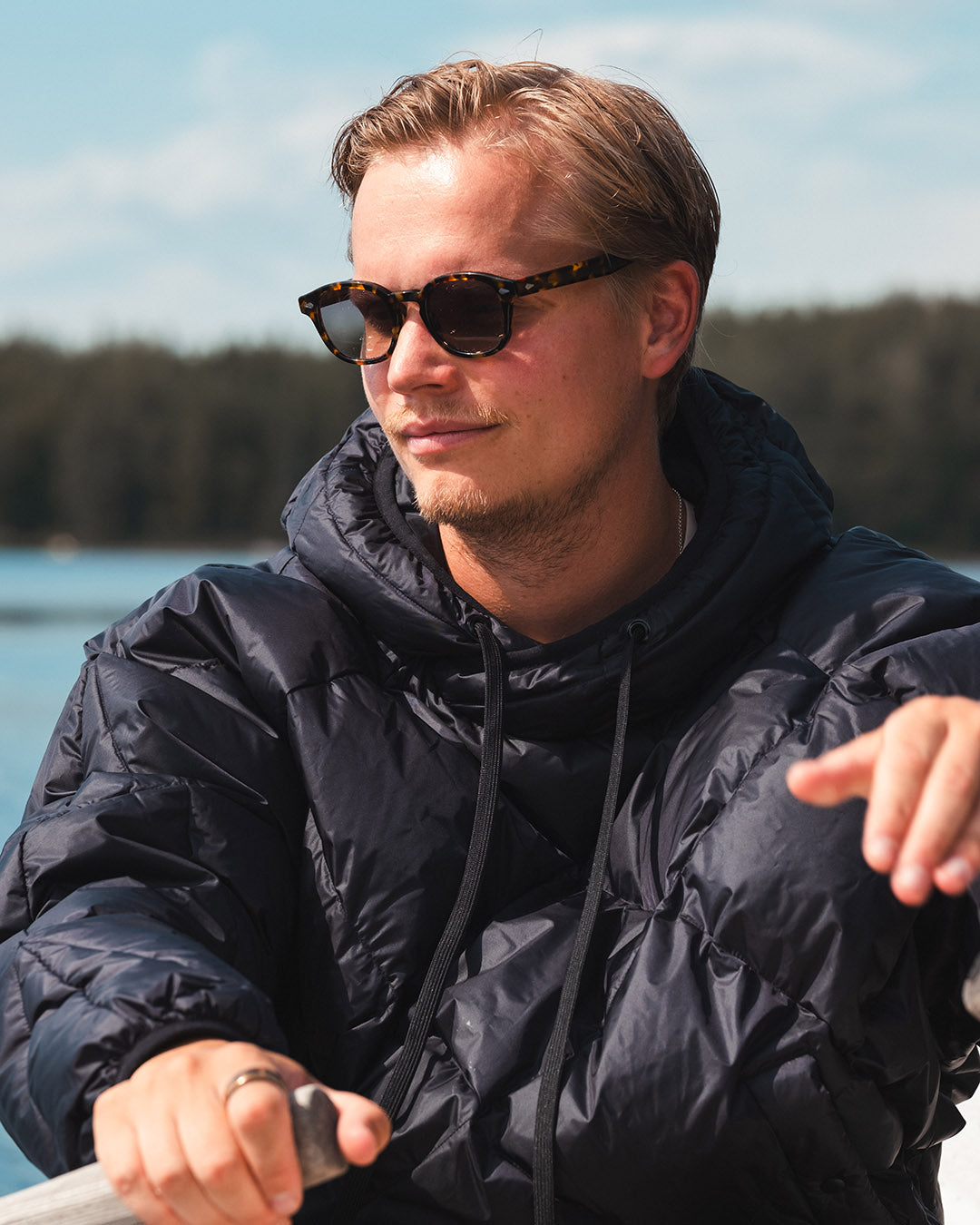 Man wearing sunglasses and a black Velho down anorak by a body of water with trees in the background