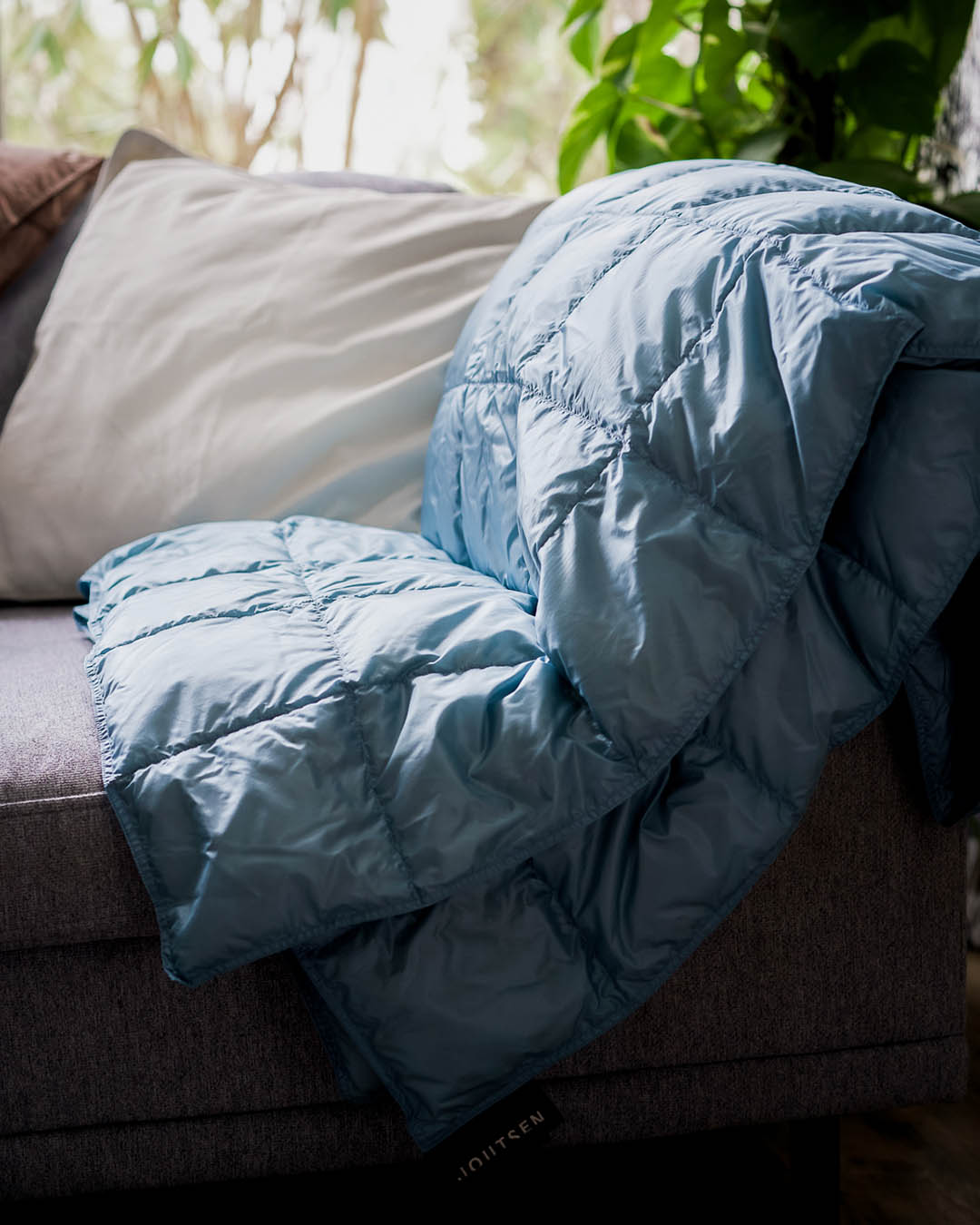 The ice blue quilted Kulkuri down blanket is draped over the armrest of a grey sofa. In the background, a light-colored pillow and green houseplants are visible, with natural light filtering in through the window behind them.