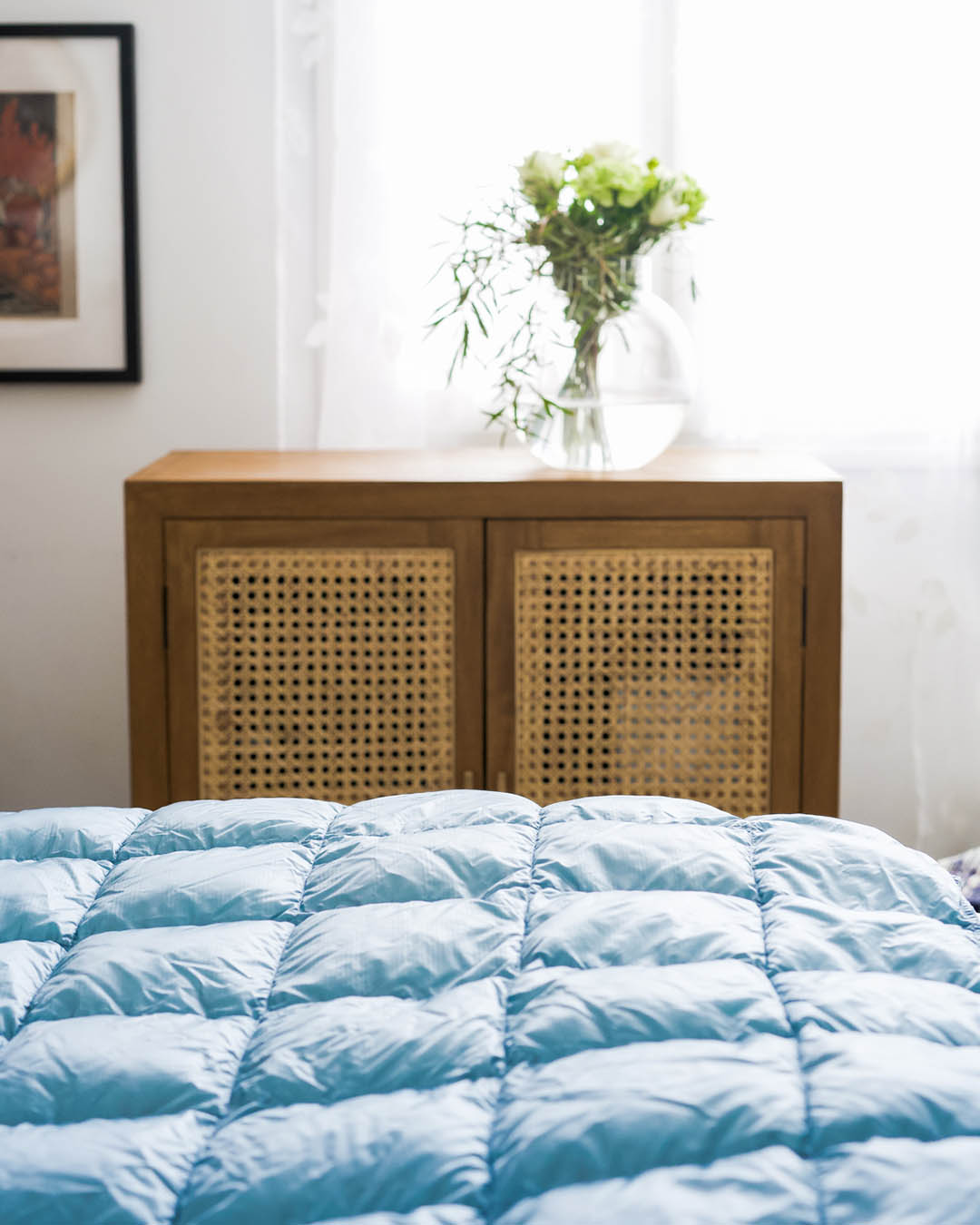 The light blue Kulkuri down blanket (color: ice blue) is spread out on the bed in a bright room. In the background, there is a wooden cabinet with woven door panels, topped with a glass vase containing green branches.