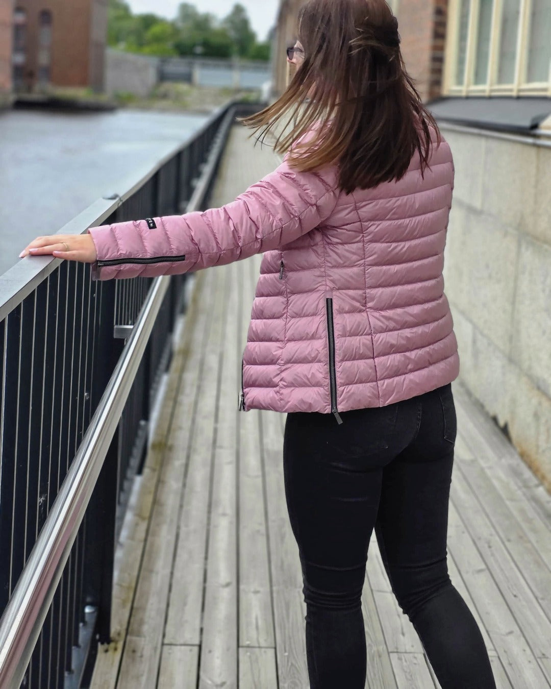 A woman stands by the water with her back to the camera, holding onto a metal railing with one hand. She is wearing a pink Maana light down coat (color: Wildflower), which has zippered slits both in the side seams and at the sleeve ends.