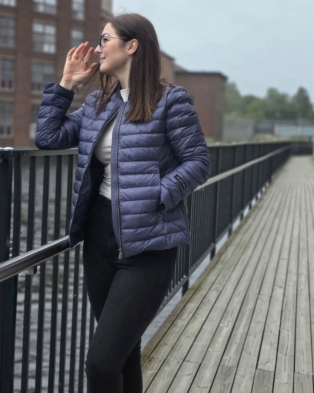 A woman leans against the metal railing of a wooden bridge by the water. She is wearing a blue Maana light down coat (color: Storm). The coat features horizontal quilting and zippered slits at the sleeve ends and side seams