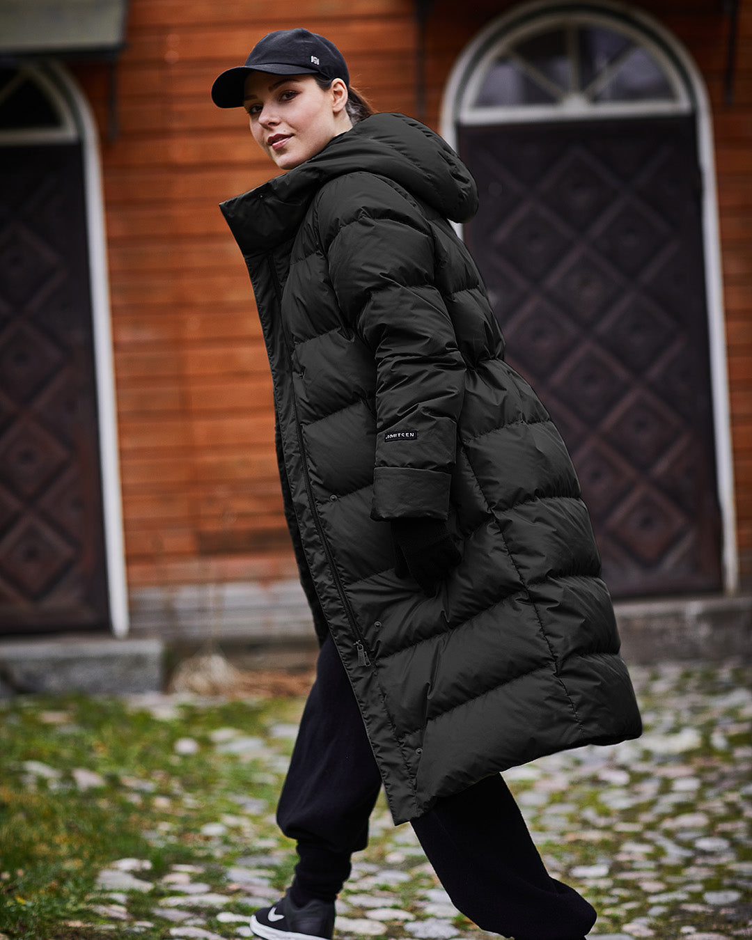 A woman walks outdoors wearing a black, horizontally quilted, long Ulrika down coat, a black cap, and black pants. In the background, there is an old wooden building and a stone-paved yard.
