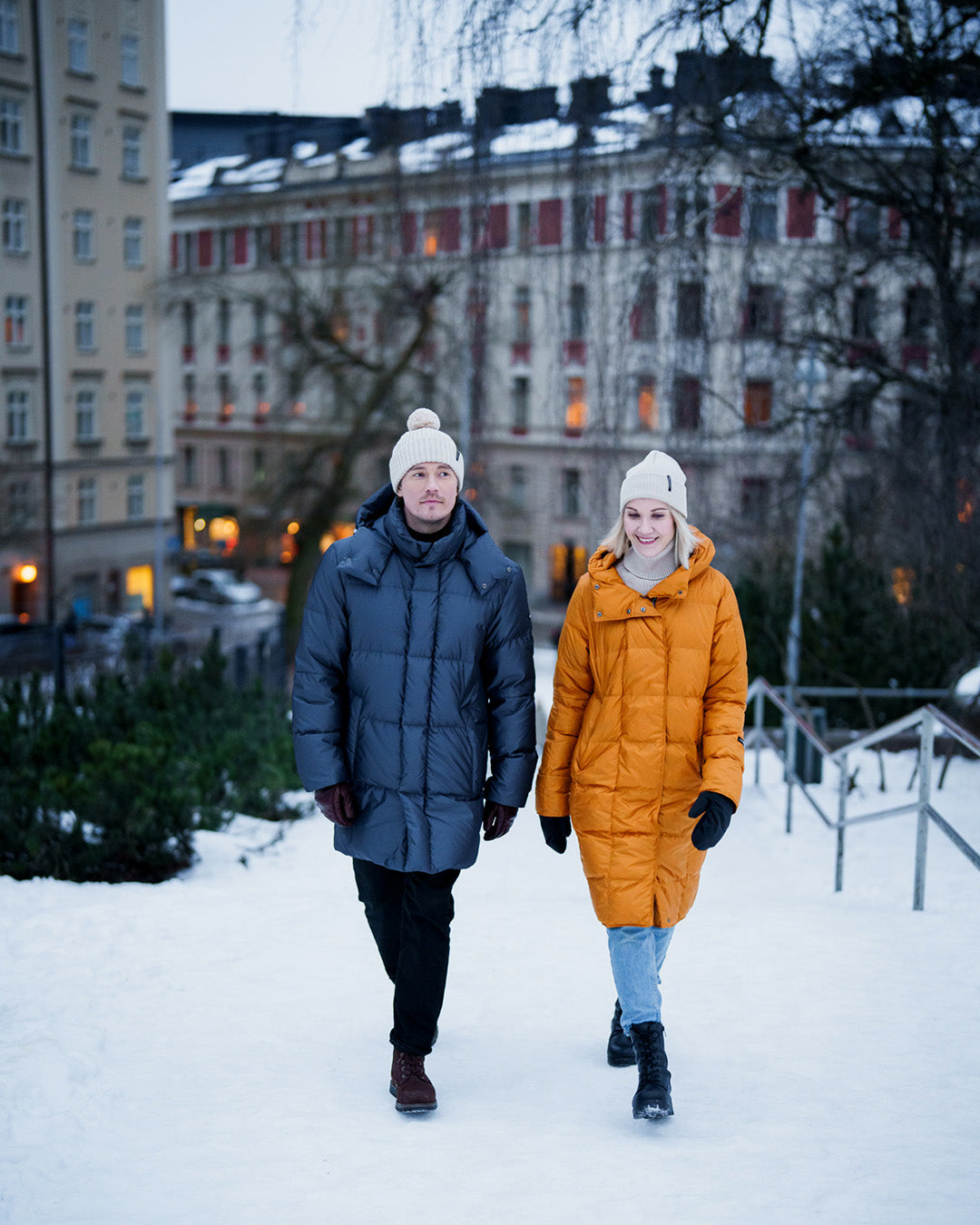 Two people walk in a snowy urban environment. One wears a yellow Essi light down coat (color: Amber), and the other wears a long men's down coat; both have hoods and square quilting. Apartment buildings and a winter landscape are visible in the background.