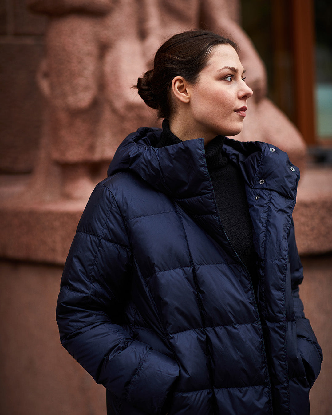 A woman stands in an urban setting wearing a dark blue, long Essi light down coat (color: Night Sky) with a hood and square quilting. The coat is partially open, revealing a black high-neck top underneath.
