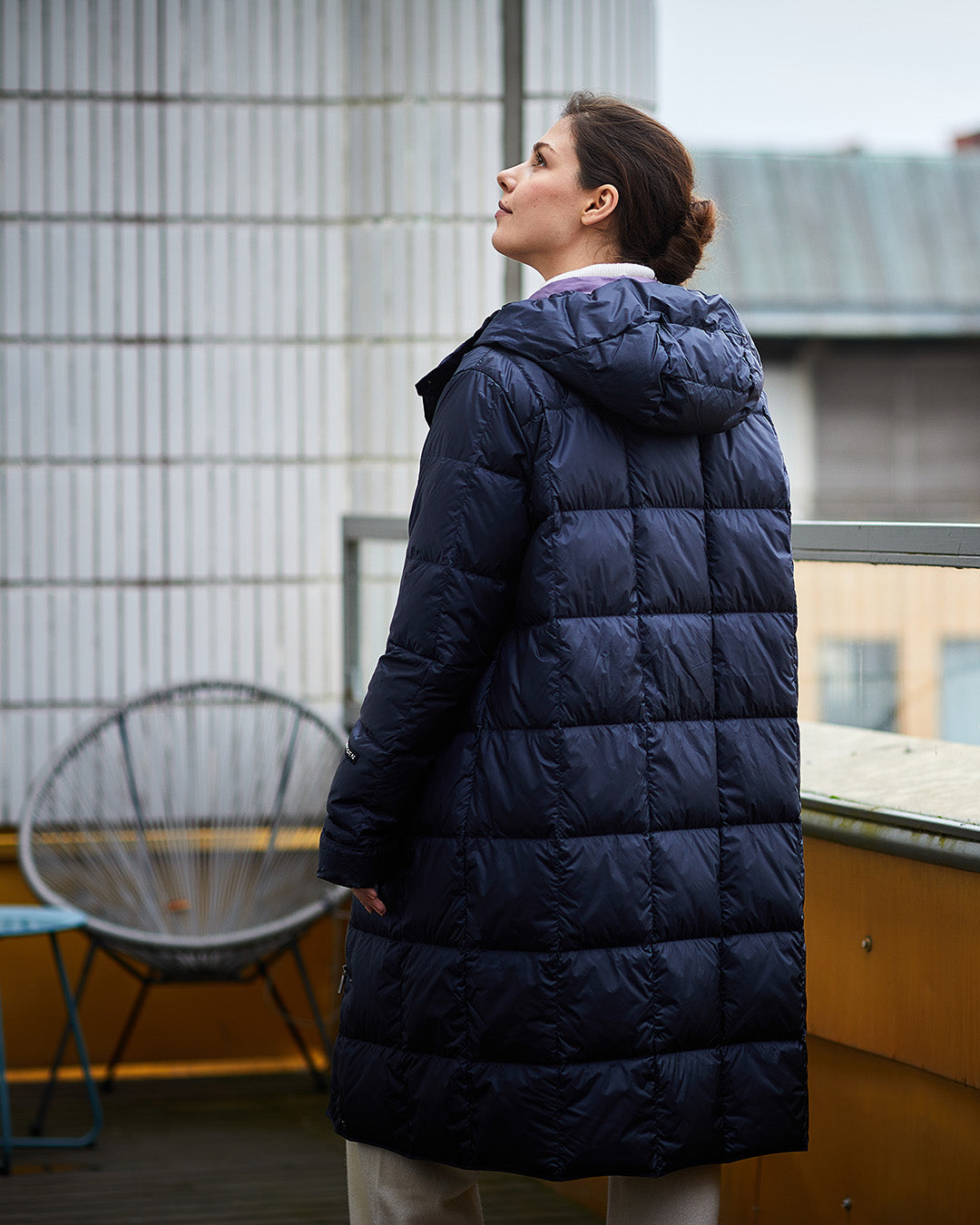 A woman stands with her back to the camera wearing a dark blue, long Essi light down coat (color: Night Sky) with a hood and square quilting. In the background, there is a balcony or terrace and some buildings.