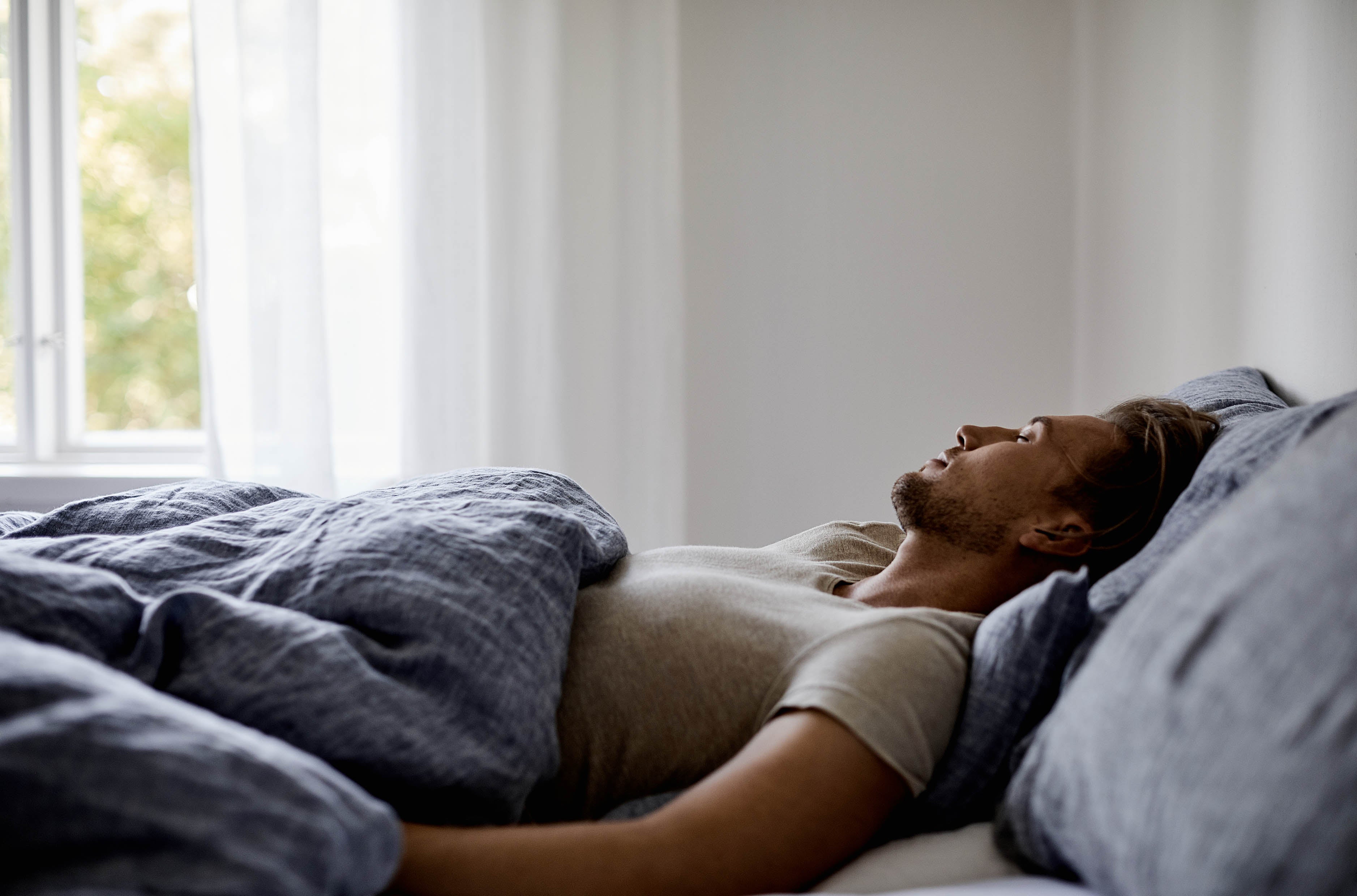 Man lying in bed on his back in a bright bedroom. White curtains covers a half of a window.