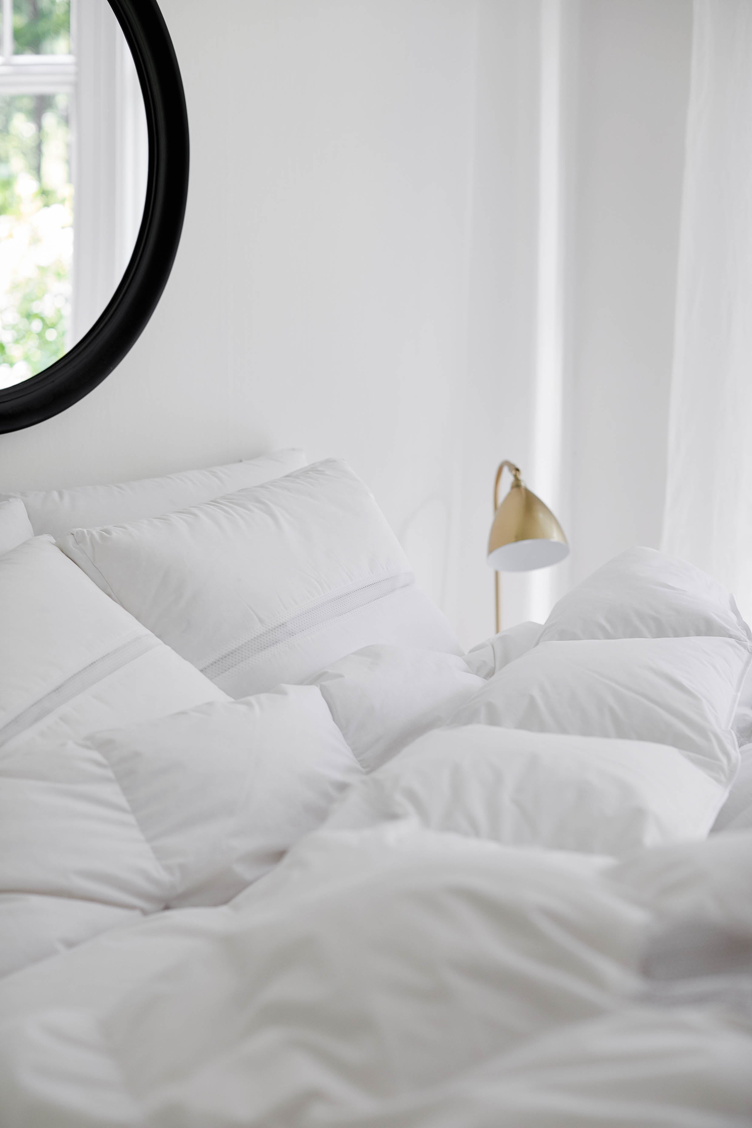 White down duvet and down pillows on a bed with a round mirror and window in the background.