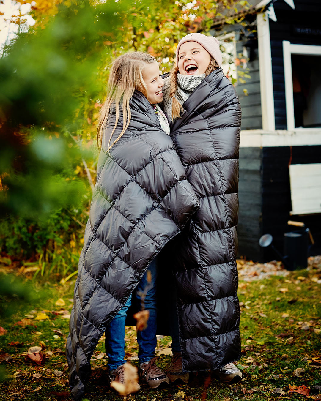 Two smiling children are standing outside in an autumnal yard, wrapped together in a dark grey Kulkuri down blanket (color: anthracite). They are laughing and looking at each other, with a cabin and colorful fall foliage visible in the background.