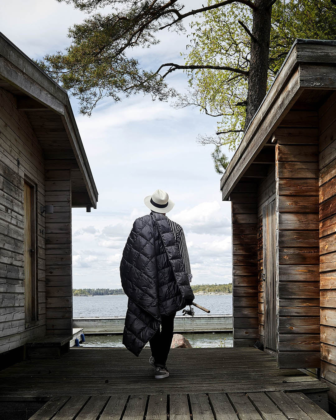 A person walks onto a dock between two log saunas, carrying a dark grey Kulkuri down blanket (color: anthracite) over their shoulder. In the background, there is a lake view, a tree, and a cloudy sky.
