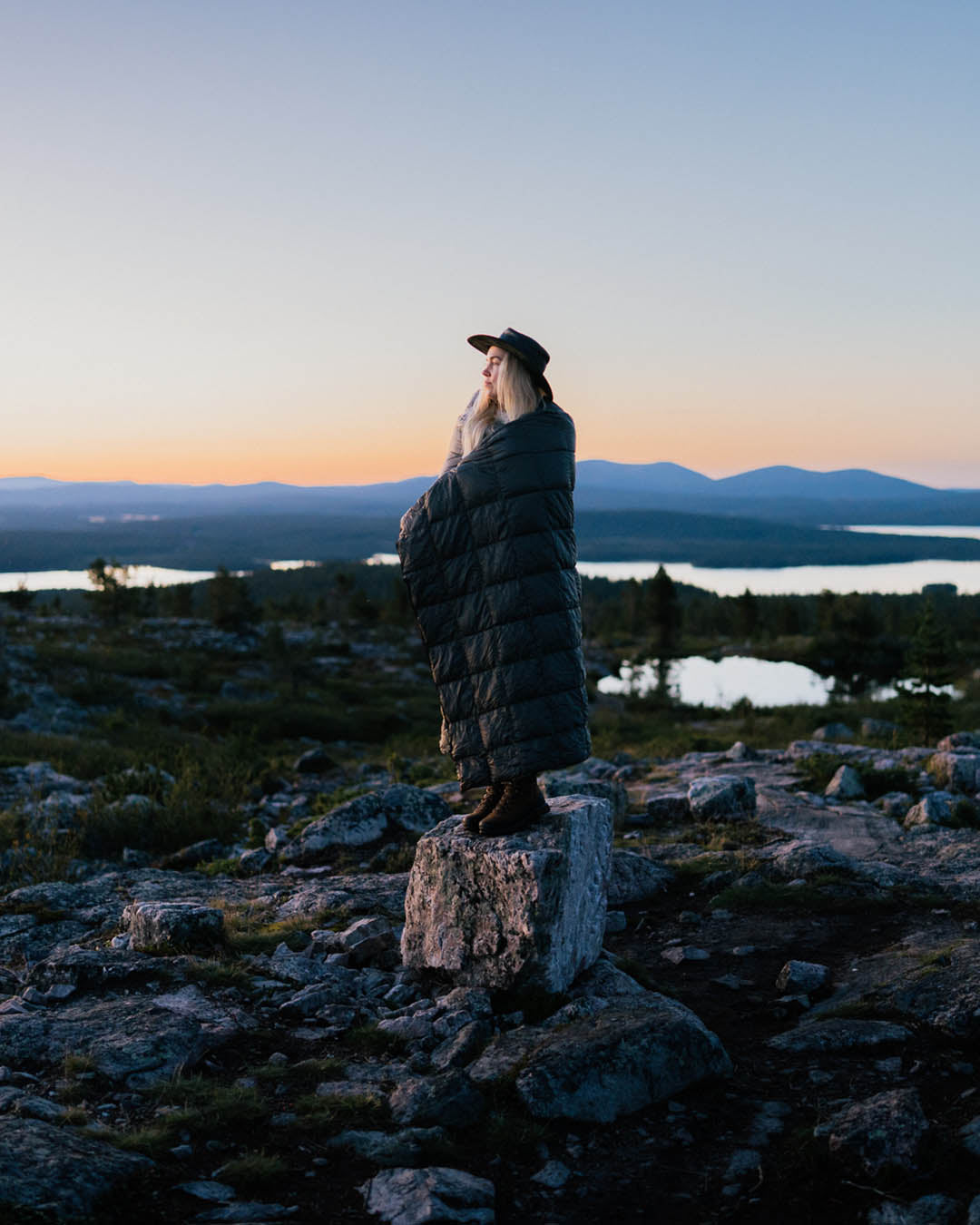 A person stands on a rock in an open fell landscape at sunset. They are wrapped in a long, quilted, dark grey Kulkuri down blanket (color: anthracite) and gaze toward the horizon.