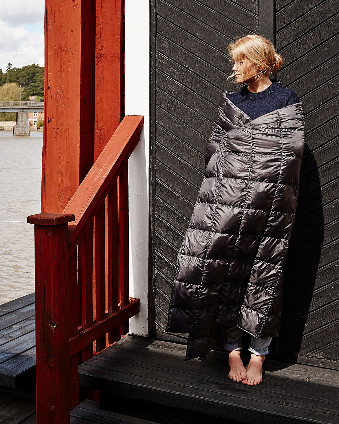 A person stands barefoot on a wooden terrace, wrapped in a dark grey Kulkuri down blanket (color: anthracite). They lean against a black plank door and look to the left. In the background, there is a river landscape and green trees on the opposite shore.