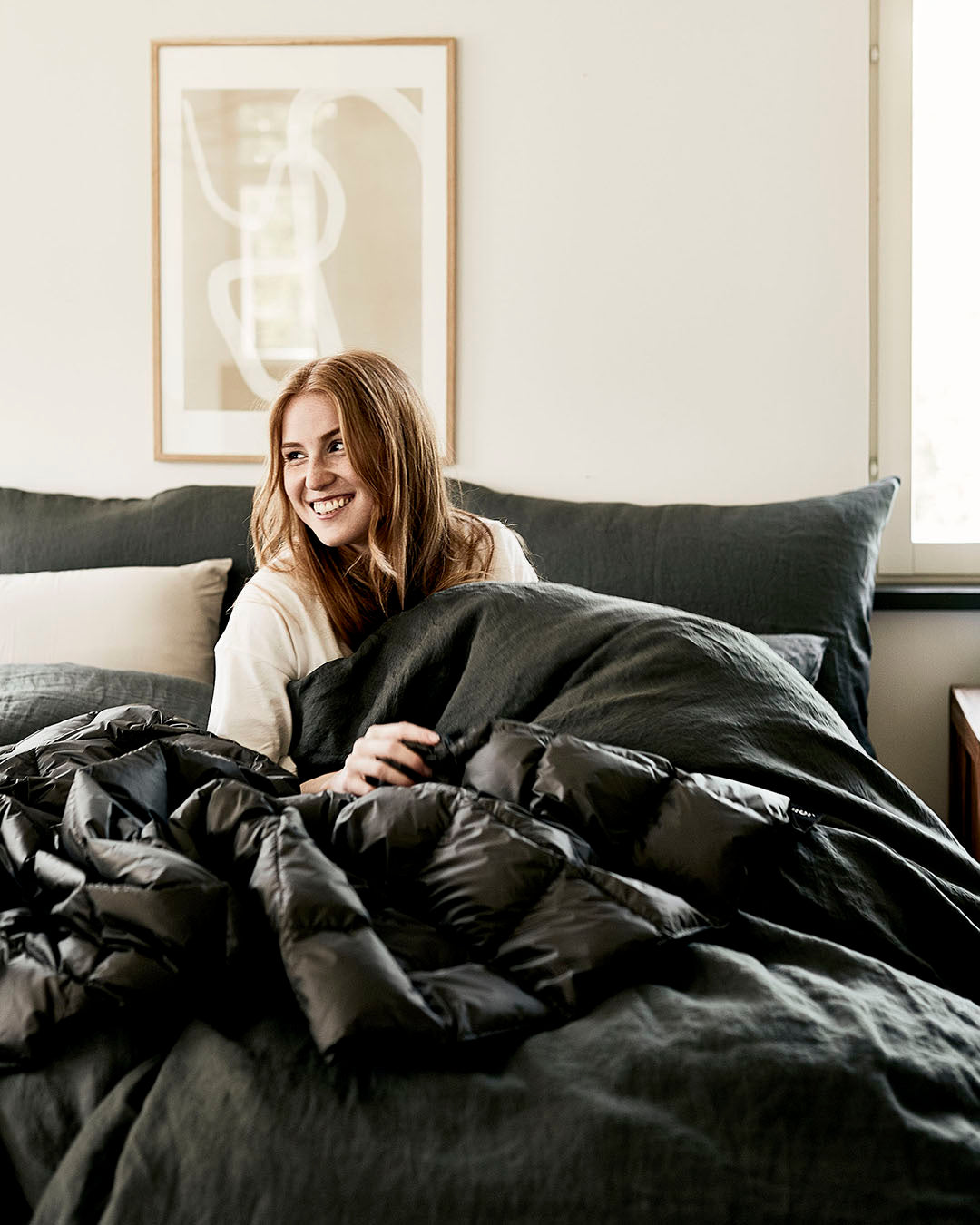 A smiling person is sitting on a bed surrounded by dark grey bedding, wrapped in a shiny, quilted Kulkuri down blanket (color: anthracite). In the background, there is a light-colored wall, a graphic art print, and a window letting natural light into the room.