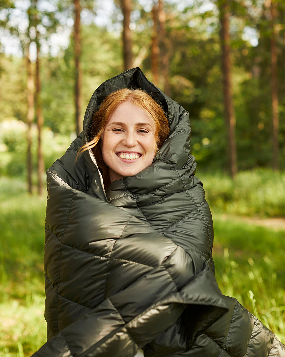 A smiling person is wrapped in a quilted olive green Kulkuri down blanket. In the background, a summer forest is visible with sunlight and lush undergrowth.