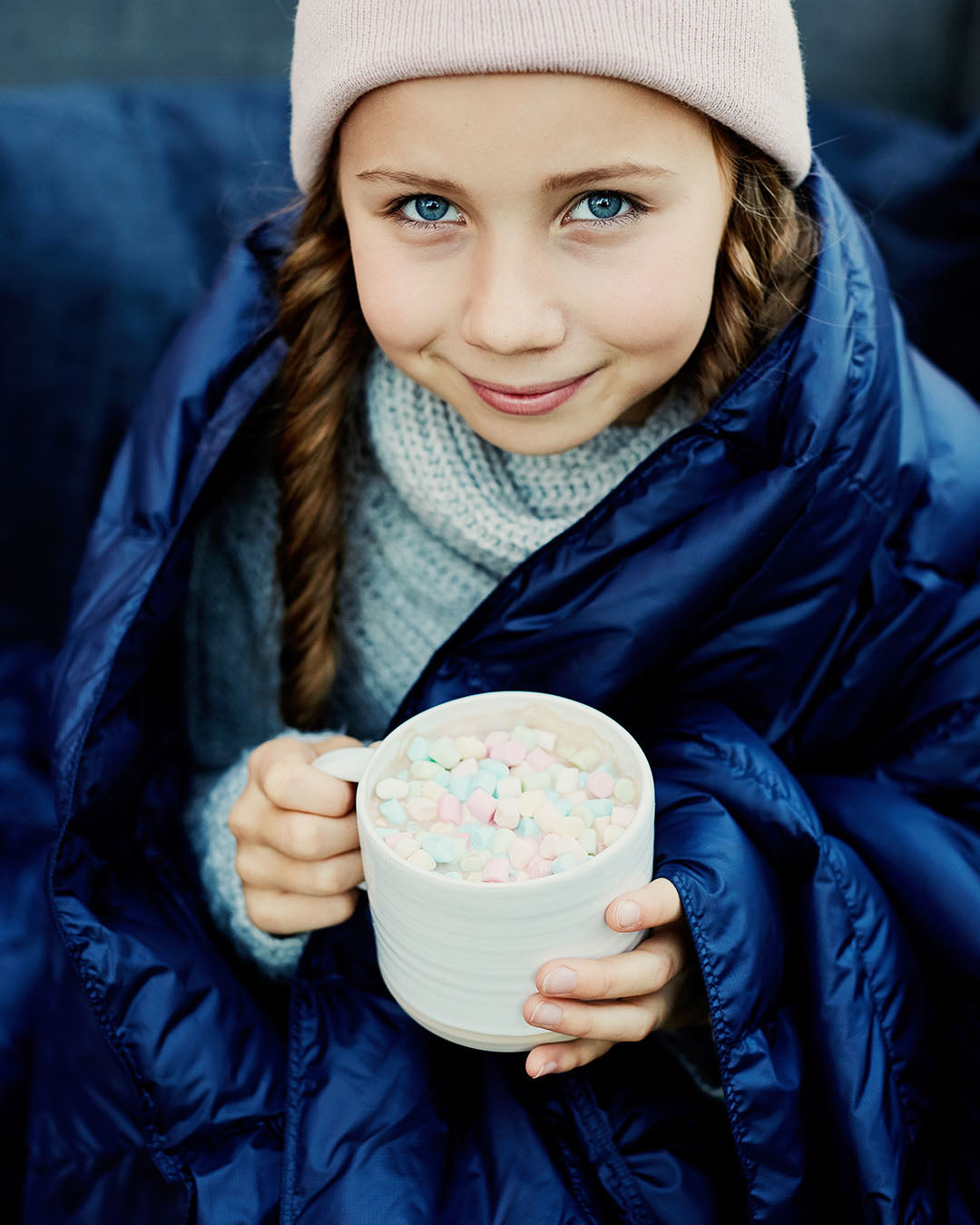 A smiling child is sitting wrapped in a dark blue Kulkuri down blanket, holding a mug filled with marshmallows. They are wearing a pink beanie, a grey wool sweater, and have braided hair.