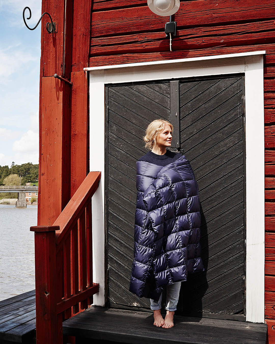 A person stands barefoot on a wooden terrace, wrapped in a dark blue quilted Kulkuri down blanket. They lean against a black plank door and look to the right. In the background, there is a river landscape and green trees on the opposite shore.