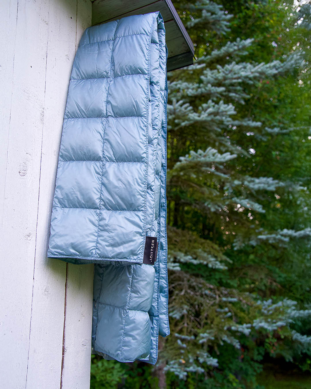The light blue Kulkuri down blanket (color: ice blue) is folded and hung on a hook attached to the wall of a white building. In the background, green forest scenery and conifer trees are visible in the summer light.