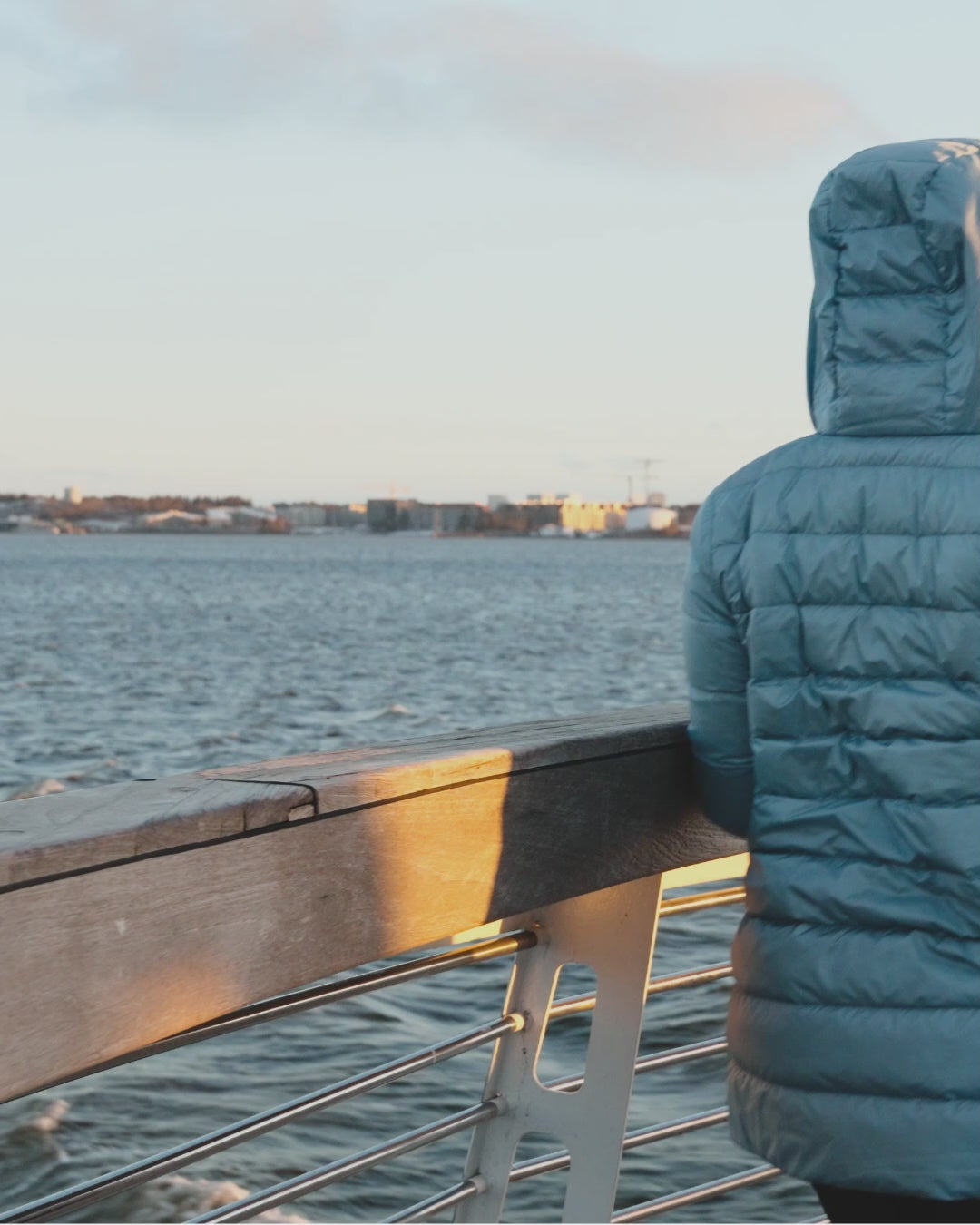 A woman walks on a pier in the evening sun, wearing a light blue Ruut light down coat (color: harebell) with horizontal quilting and a hood. She has a chunky brown beanie on her head and black mittens on her hands.