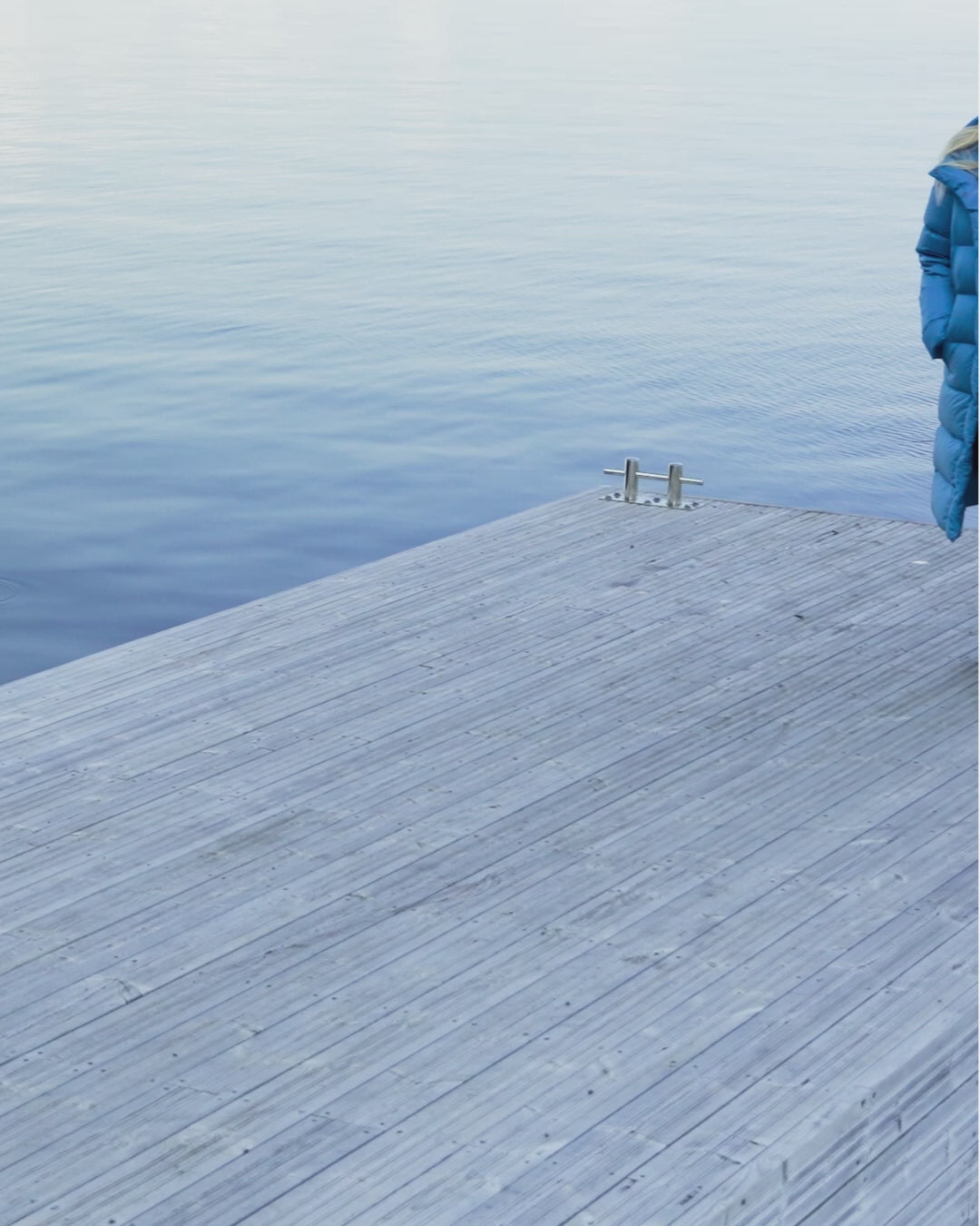 A blonde woman walking on a large pier wearing a blue (color name: Blue butterfly) long down coat. The coat’s zipper is open, and she has her hands in the coat pockets. She takes a few steps with a smile on her face.