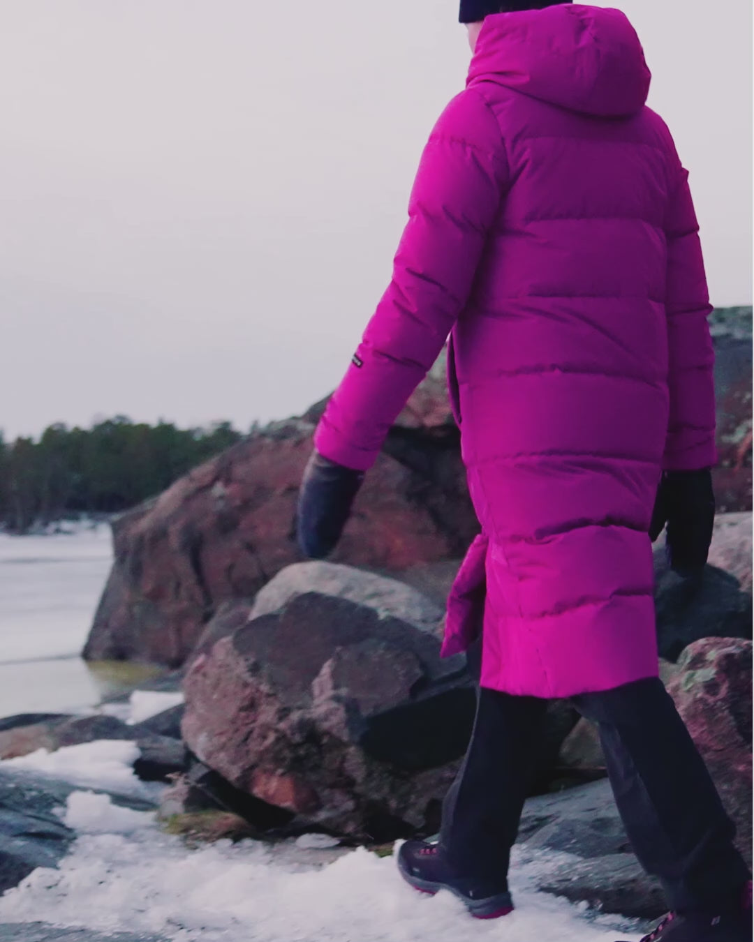 A woman is walking on a rocky shore in a winter landscape, wearing a horizontally quilted, fuchsia-colored Ulrika down coat (color: peony). She is also wearing a black beanie, black outdoor trousers, and hiking boots.