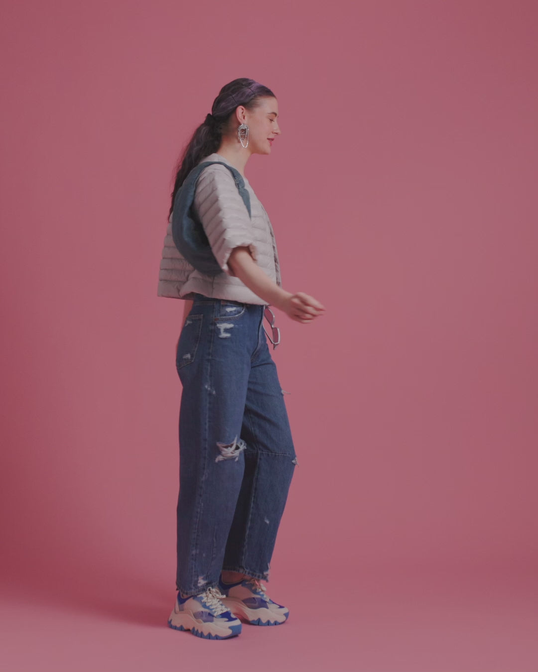 A woman is dancing in front of a backdrop, wearing a light-colored, short Lupiini light down jacket (color: sugar cloud) and ripped blue jeans. She holds a dark handbag on one shoulder.
