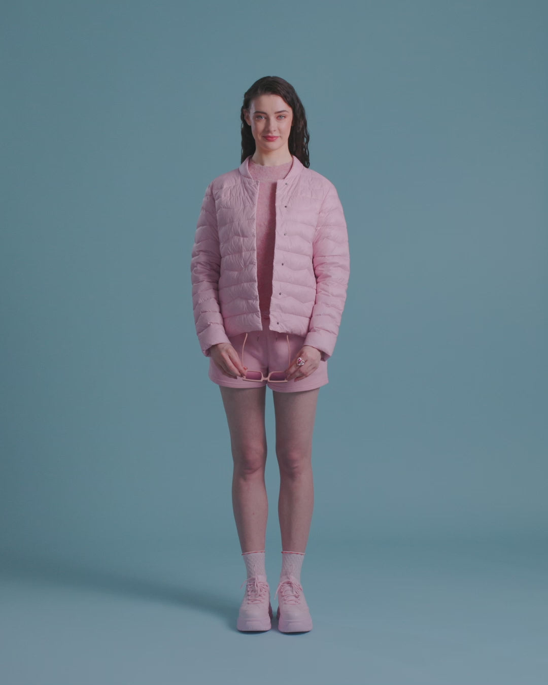 A woman dressed in a pink Lootus light down jacket (color: strawberry mousse), a matching knit sweater and shorts. The outfit includes pink sneakers and sunglasses. She is putting the sunglasses on her head and rolling up her sleeves.