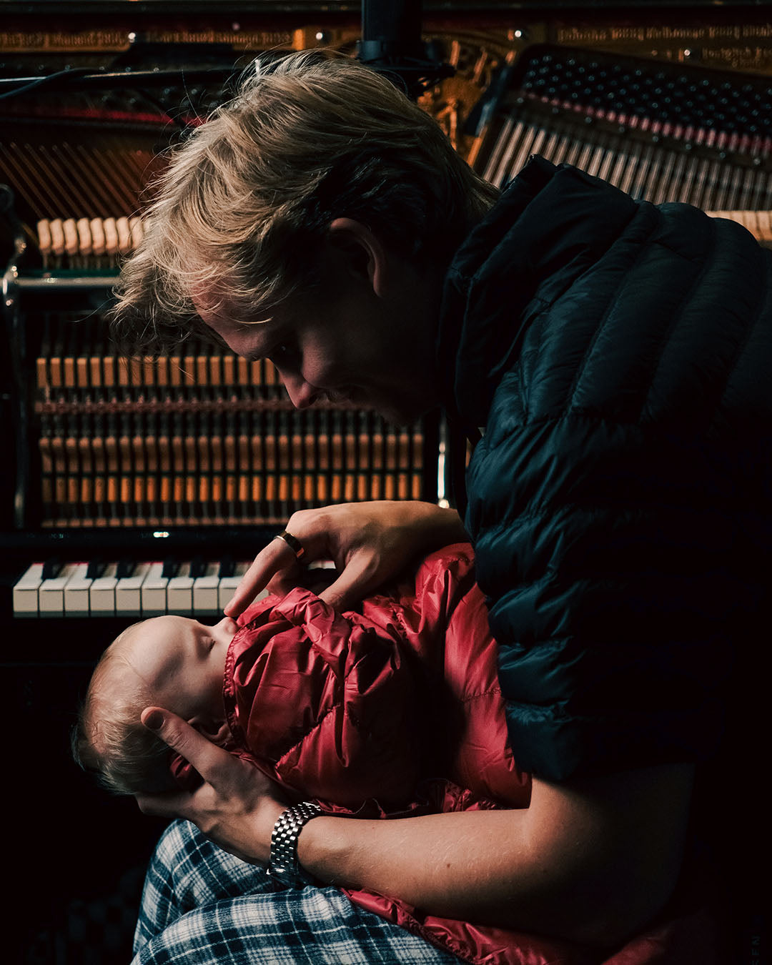 Person holding a child in red Kulkuri down blanket in front of a piano.