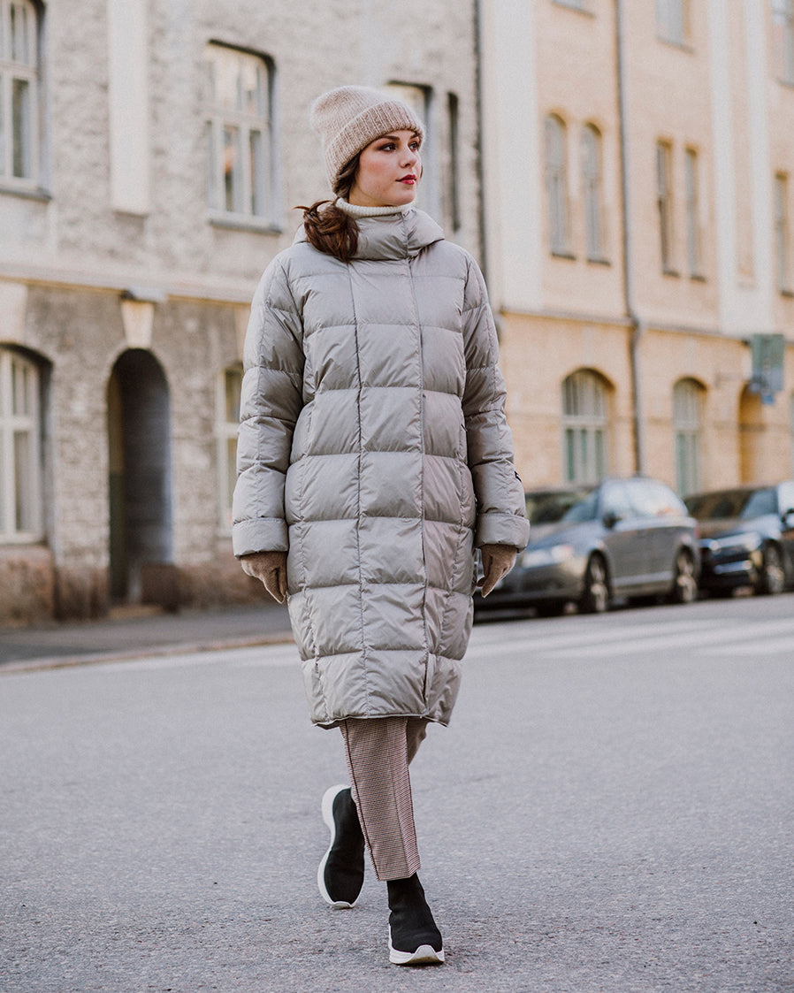 A woman walks along a city street wearing a light-colored, long Essi light down coat (color: champagne) with square quilting and a high collar. Her outfit also includes a beige beanie, striped pants, and black shoes with white soles.