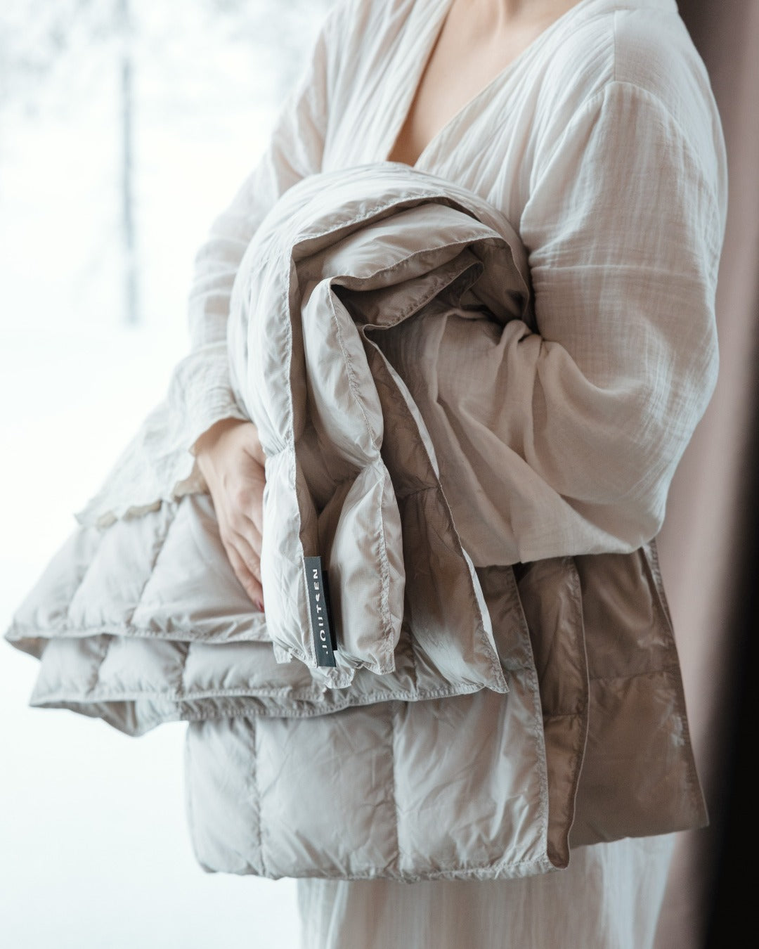 Folded beige (color:seashell) down blanket with a brand label held by a person wearing a white outfit.