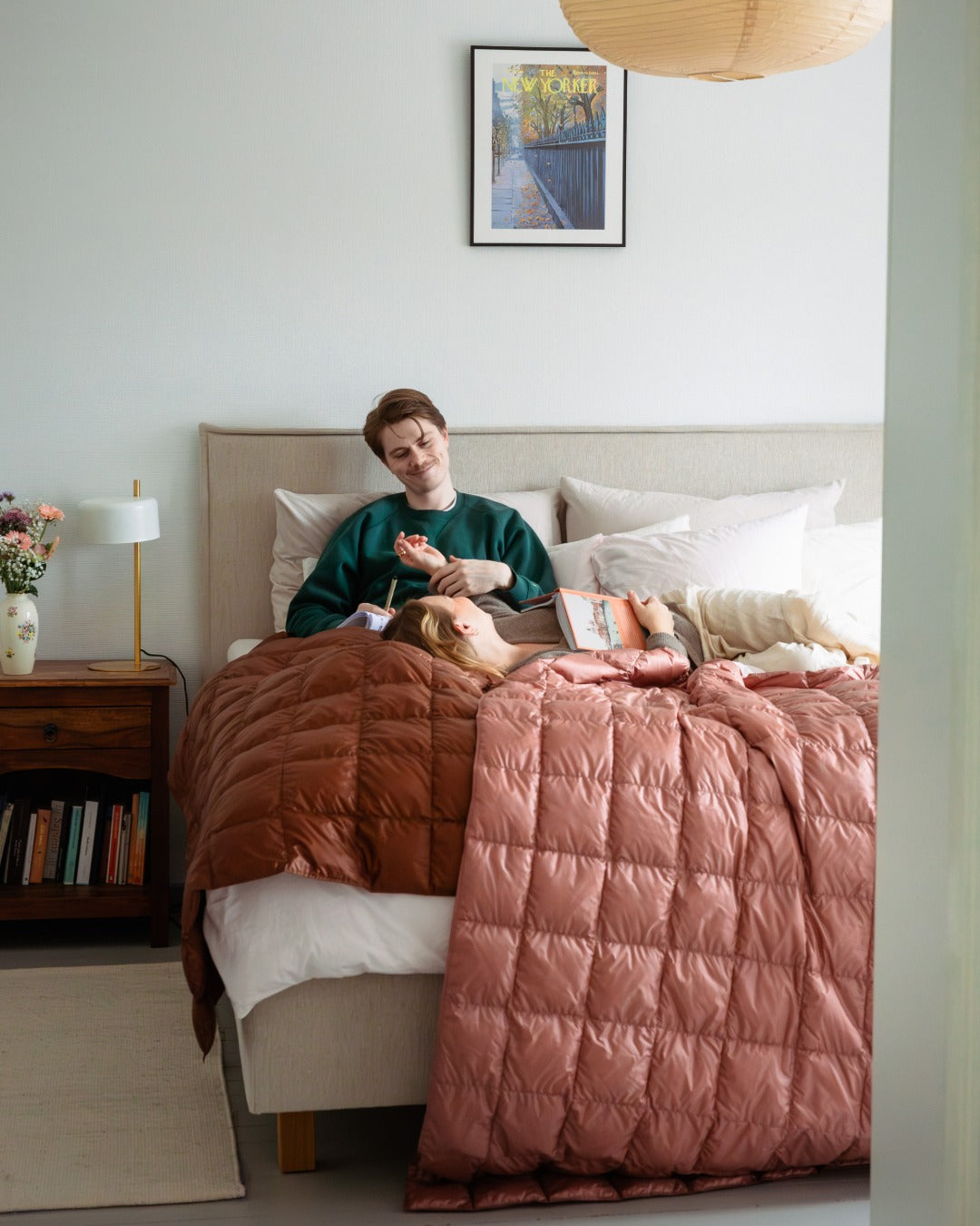 Person sitting on a bed with a light pink (color: old rose) and brown (color: chocolate mousse) down blankets, surrounded by books and a laptop.
