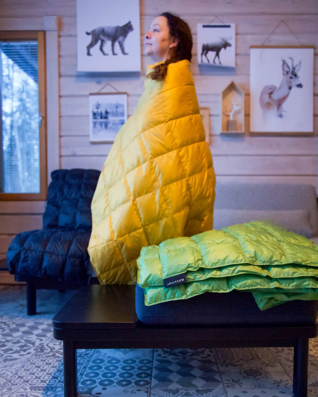 Person wrapped in a yellow Kulkuri down blanket in a cozy room with wooden walls and framed pictures.
