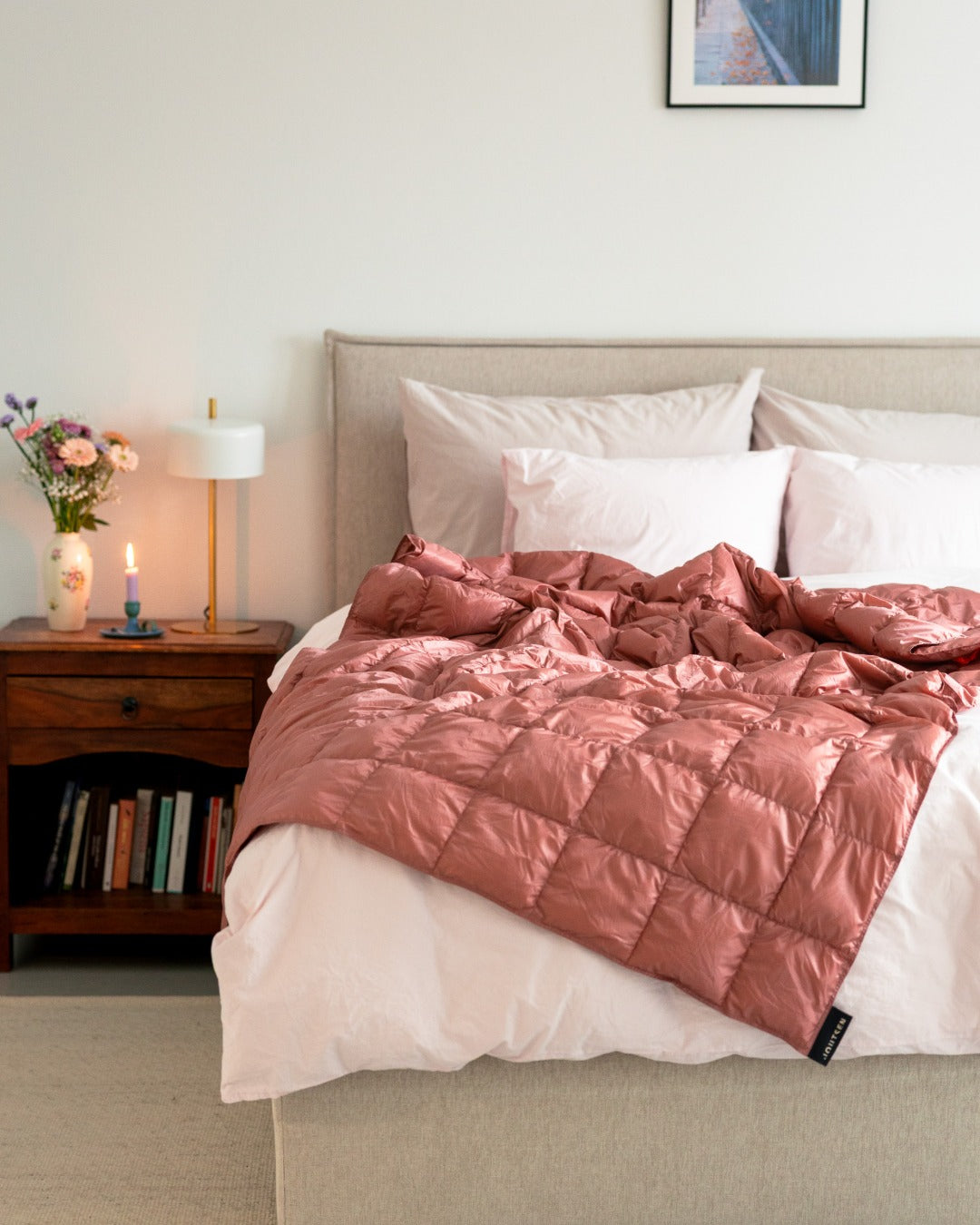 Bedroom with a bed featuring a light pink (color: old rose) Kulkuri down blanket, nightstand with books and lamp, and wall art.