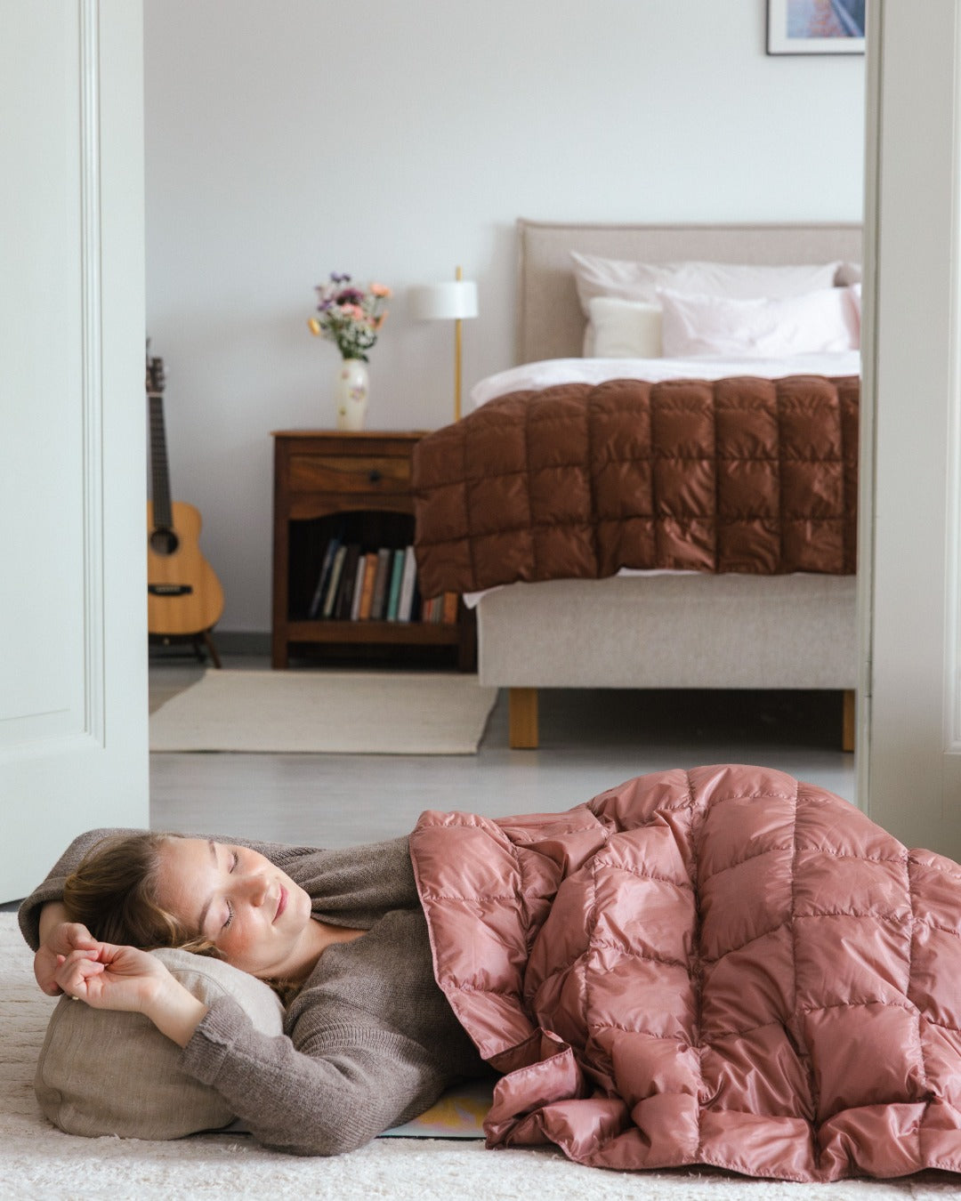 Woman lying on a floor with a light pink (color: old rose) Kulkuri down blanket in a bedroom. There is a bed in the backround with brown (color: chocolate mousse) Kulkuri down blanket.