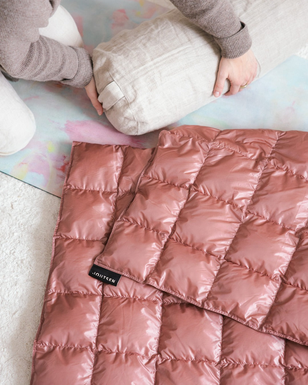 A light pink (color: old rose) Kulkuri down blanket on a colorful surface with a hand reaching towards it.
