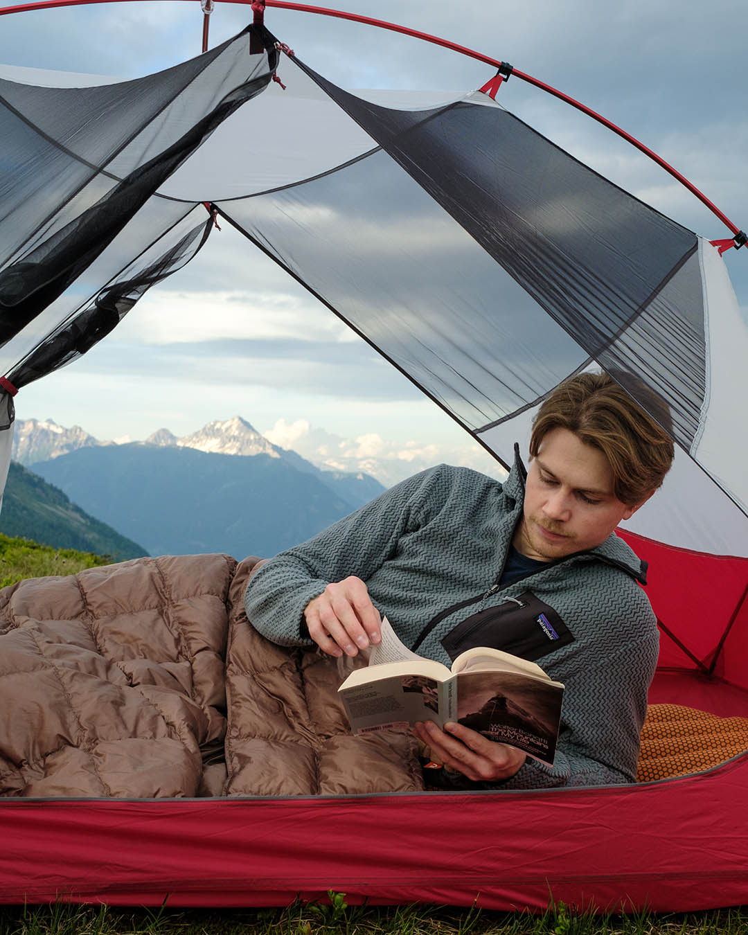 A man lies under a brown (color: hazelnut) Kulkuri down blanket with square quilting, inside an open tent in a mountain landscape, reading a book. He is wearing a grey fleece jacket, and in the background, snow-capped mountains and a cloudy sky are visible.