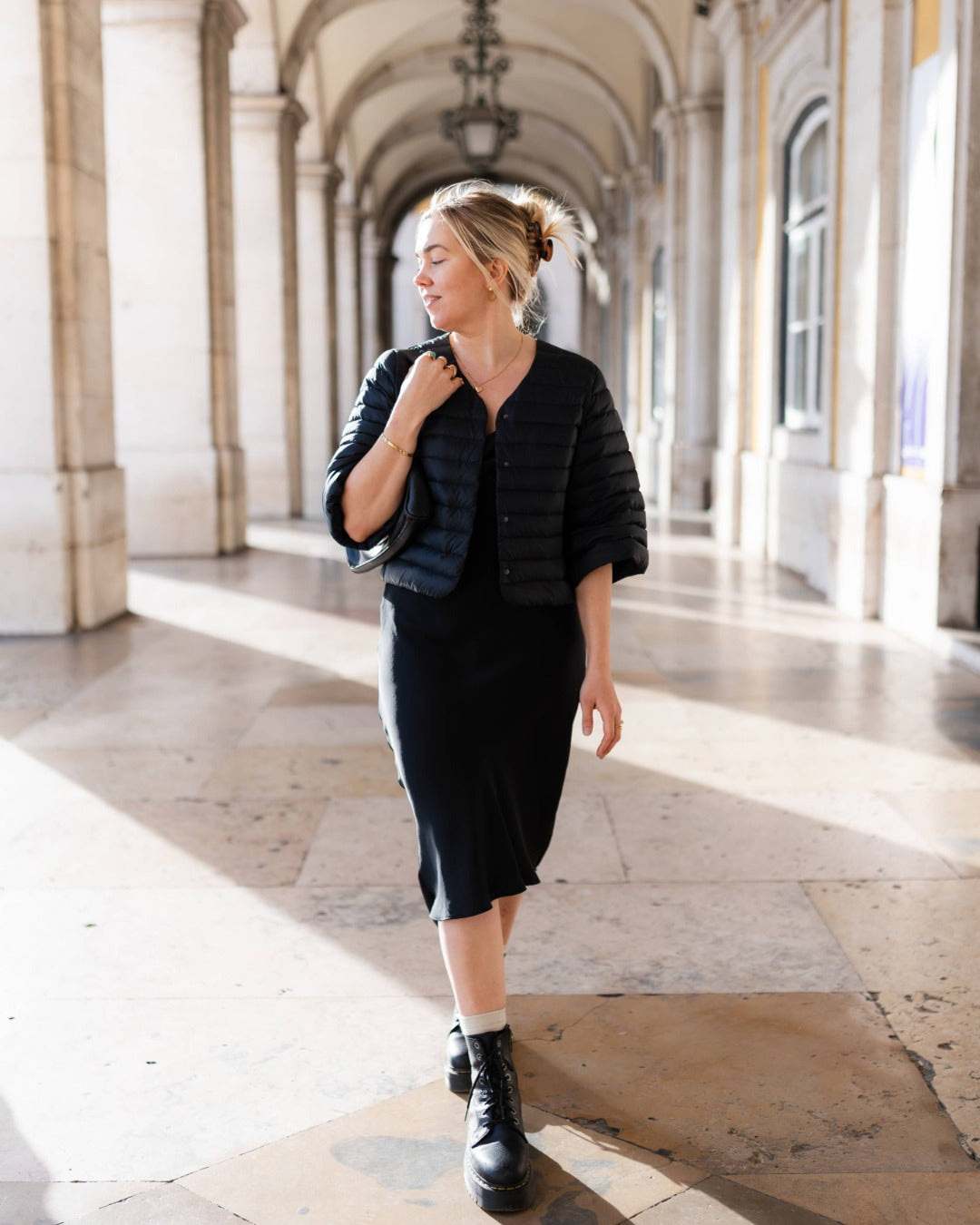 Woman in a black down bolero walking through a sunlit architectural hallway