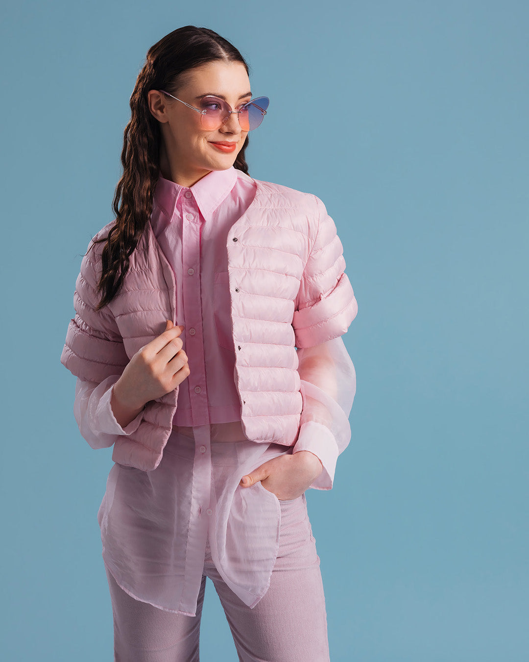 A woman dressed in a horizontally quilted light pink, short Lupiini light down jacket (color: strawberry mousse), layered over a pink collared shirt and a sheer long blouse. The outfit is completed with light-colored trousers and purple sunglasses.