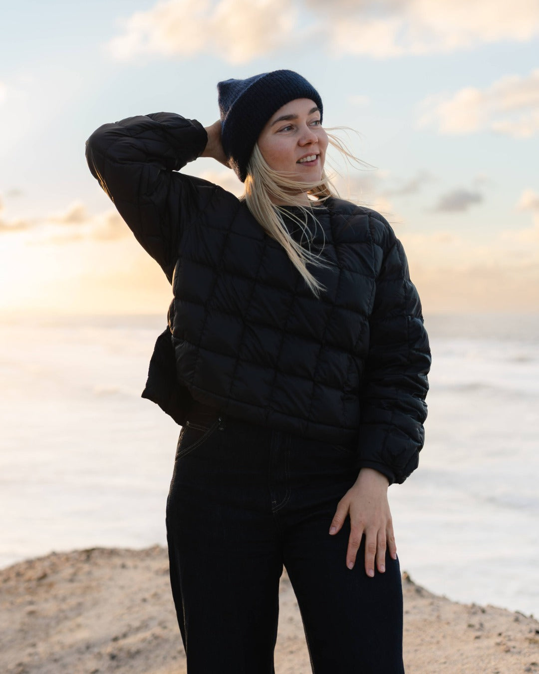 Person wearing a black down college and beanie standing on a beach with sunset in the background